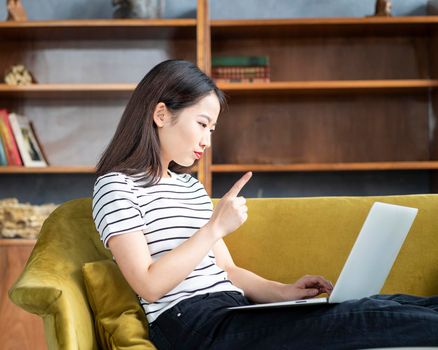 Asian woman finger wagging looking at laptop, working at home, liying on sofa. Beautiful Japanese female scolding, educating child