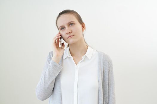 Calm, unemotional woman talks on phone, listens to other person, does not react. Quiet girl listens carefully using mobile phone. White gray wall, copy space