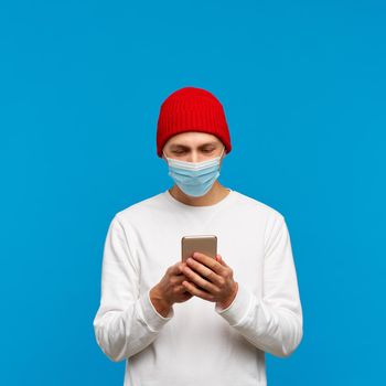 Portrait of man with medical protective face mask, texting on mobile phone. Male write message isolated on bright yellow colored background. White jumper and red hat.