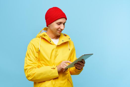 Portrait of positive cool man in warm autumn top outdoor clothes holding tablet, male isolated over blue color background. Bright casual wear, yellow cloak