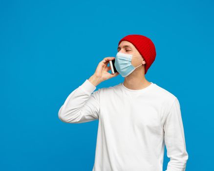 Portrait of man with medical protective face mask, speaking to mobile phone. Male isolated on bright yellow colored background. White jumper and red hat.