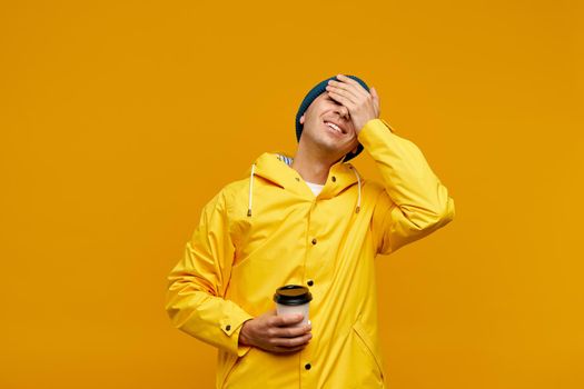 Portrait of positive cool man in warm autumn top outdoor clothes holding coffee and closing eyes with hands, male isolated over yellow color background. Bright casual wear, yellow cloak