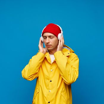 Portrait of positive cool man holding headphone listening music with closed eyes, male isolated over blue color background. Bright casual wear, yellow cloak and red hat