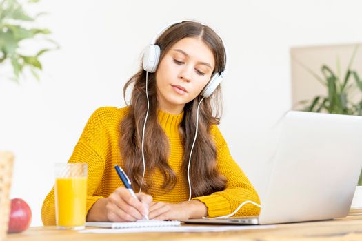 Happy female in wireless headphones studying online course, using pc and writing in notepad, copy space. Gig economy, digital nomad, distance education. Side view of mature woman work from home