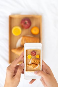 Blogger taking photos of food, shooting Breakfast in bed at hotel on mobile phone, tray with juice, fruit and croissant on white sheets