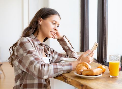 Woman looking for information on Internet, smiling and using mobile, enjoying breakfast. Concept for digital addiction, morning routine. Sunday slow life