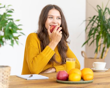 Young girl bitting red apple, smiling. Beautiful woman with long hair in bright yellow sweater eating fruits for detox