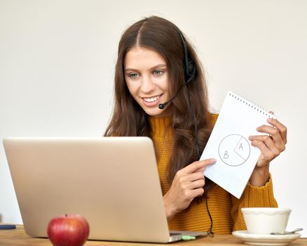 Happy woman in headset speaking by conference call and video chat on laptop in office. Explaining and showing diagram in notebook. Online education, course. Support service agent consulting customer.