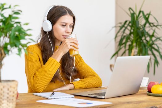 Young girl with headphones talking on conference calls,drinking orange juice, smiling. Beautiful woman with long hair in bright yellow sweater works remotely from home