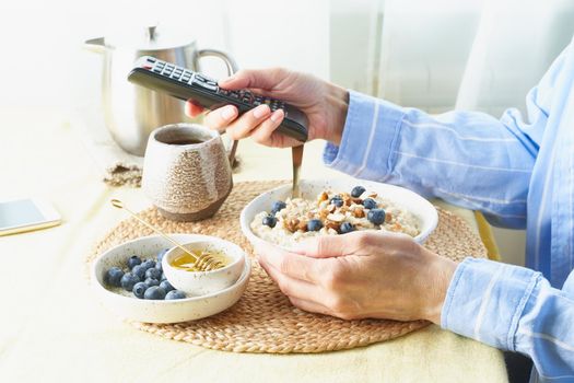 Woman is looking for information on TV, using remote control, during eating breakfast. Concept for digital addiction.