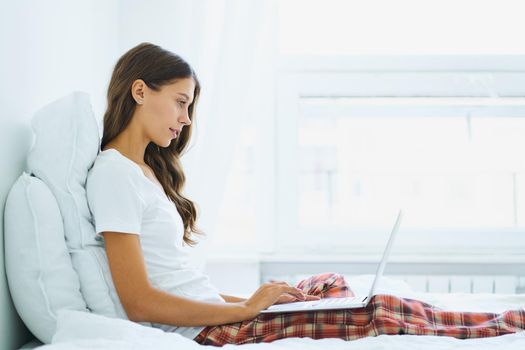 Side view of young woman sitting on bed in bedroom and typing on laptop. Work from home, distance learning for students, surfing Internet in morning after waking up