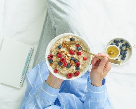 Top view faceless woman holding plate of oatmeal, sitting in bed early morning, quiet home environment. Healthy breakfast