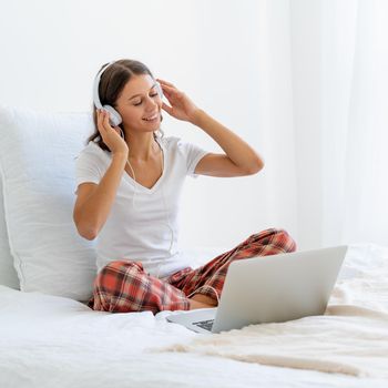 Young smiling woman sitting on bed in bedroom and listening to music or watching movie on laptop. Distance learning, surfing Internet in morning after waking up