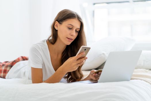 Young woman shopping online, entering code from SMS message on mobile phone, security. Female sitting on bed in bedroom and working from home on laptop