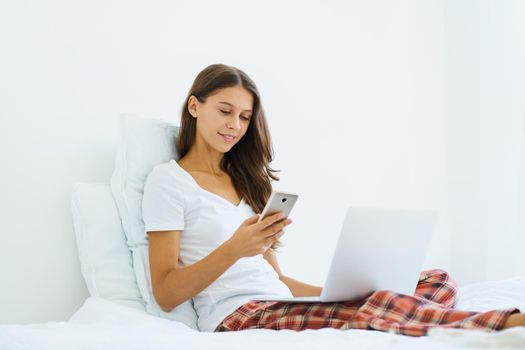 Young woman shopping online, entering code from SMS message on mobile phone, online shopping security. Female sitting on bed in bedroom and working from home on laptop