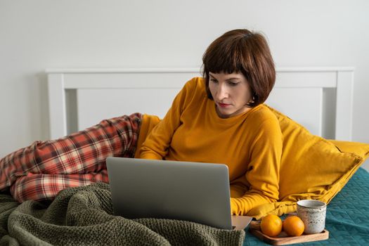 Mature woman laying on bed in bedroom and watching movie on laptop. Work from home, distance learning, surfing Internet in morning after waking up.