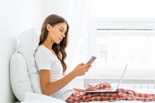 Young woman reading message from SMS on mobile phone, online shopping, work from home. Female sitting on bed in bedroom and typing on laptop