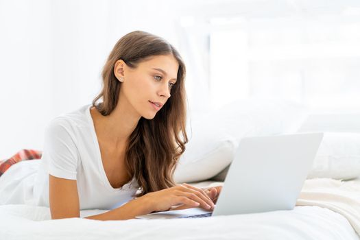 Young woman working at home laying on bed in pajamas, shopping online. Female in bedroom typing on laptop, distance learning for students, surfing Internet in morning after waking up