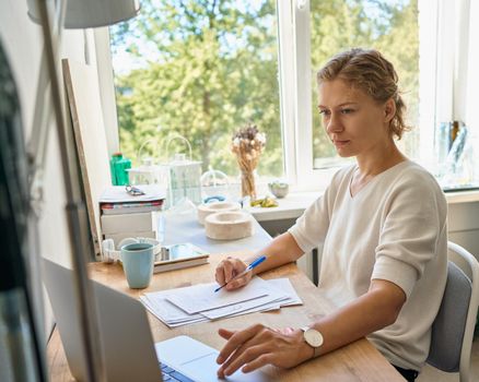 Work from home. Young female entrepreneur designer writing on paper sheet, looking at laptop, serious woman thinking and planning, happy lady in sunny day in workshop. Owner of small business