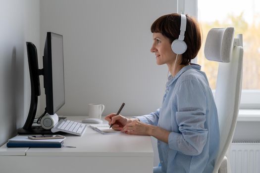 Happy female in wireless headphones studying online course, using pc and writing in notepad, copy space. Gig economy, digital nomad, distance education. Side view of mature woman work from home