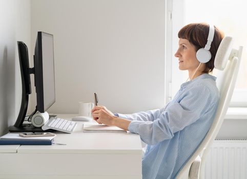 Side view of middle aged woman working remotely from home. Happy female in wireless headphones studying online course, using pc and writing in notepad, copy space.