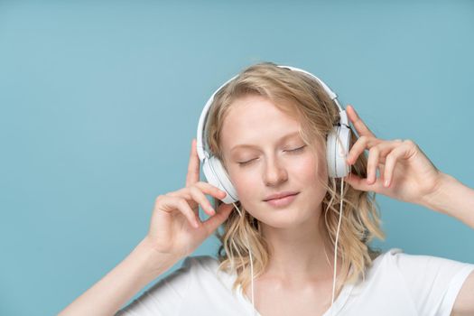 Close up Portrait of young woman closed eyes listening music via headphones on color neutral tone Aqua Menthe wall. Pretty serious clever blonde woman with curly hair in white t shirt