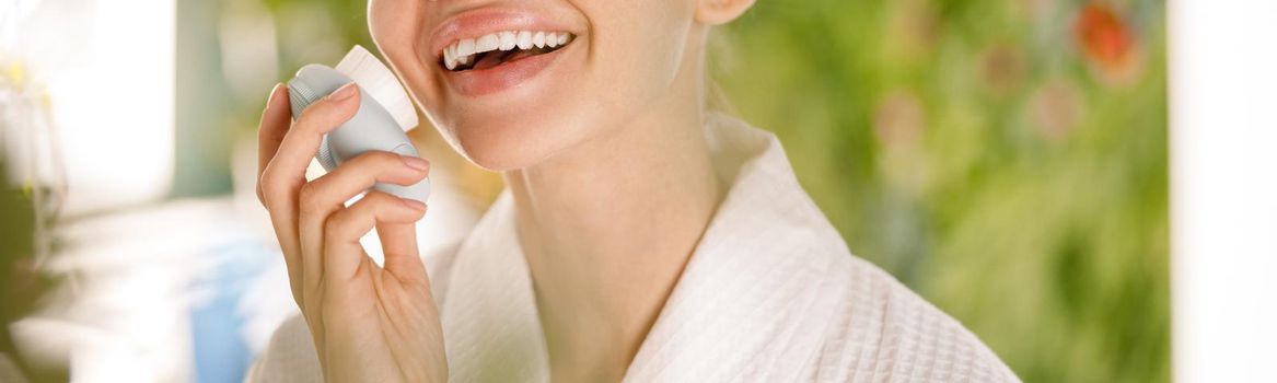 Portrait of lovely young woman with flawless skin wearing bathrobe smiling at camera while using silicone face brush for facial cleansing in the morning. Beauty, skincare, spa concept