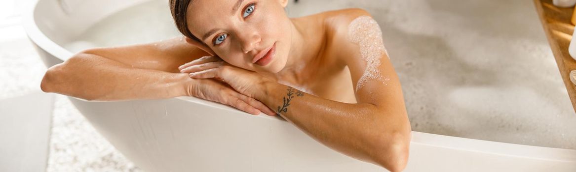 Attractive young woman feeling tired, leaning on bathtub side, bathing while relaxing at spa resort. Wellness, beauty and care concept