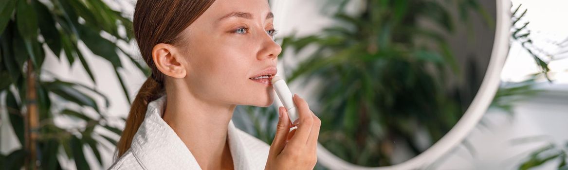 Portrait of young woman with beautiful face applying lip balm, standing in the bathroom decorated with plants. Skincare cosmetics concept