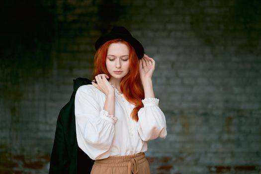 Redhead woman is resting against dark grunge wall. Female in black hat and jacket, eyes closed. Model with long thick hair. Lady posing on dark grey background looking at camera