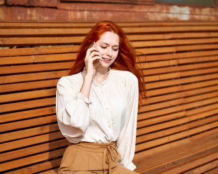 Red-haired woman talking on cell phone. Beautiful stylish fashion model sitting on bright orange bench in sun on summer day