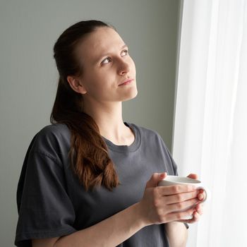 Social media, digital detox. Quarantine, self-isolation, sociophobia. Beautiful girl thoughtfully looks out window. Woman with long hair is deep in thought.