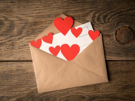 card and red hearts in open envelope from brown Kraft paper on wooden aged vintage background. Greetings with Valentine's day