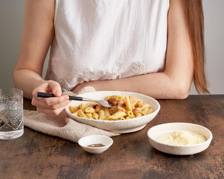 Carbonara pasta. Woman sits at table and eats pasta penne with pancetta, holding fork. Unrecognizable female in white dress. Rustic style