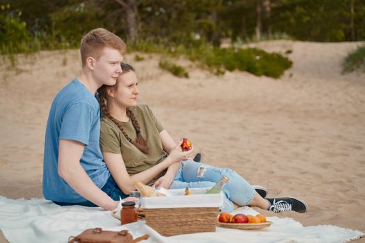 Boyfriend and girlfriend sitting on blanket embracing eating fruits looking away and smiling during picnic lovestory on beach