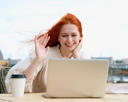 Smiling young redhead female sitting at table in outdoors cafe and chatting via video link while drinking coffee in plustic cup