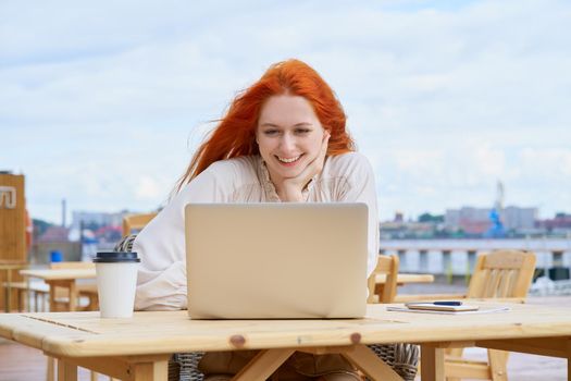 Freelancer woman sitting in cafe on street and remotely working on laptop. Female student listening courses, online education or chatting. Redhaired businesswoman typing on computer.