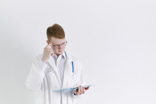 Young trainee doctor intern stands on white background. Handsome red- haired man, thinking, rubs his forehead with finger. Clipboard in his hand, stethoscope around his neck .