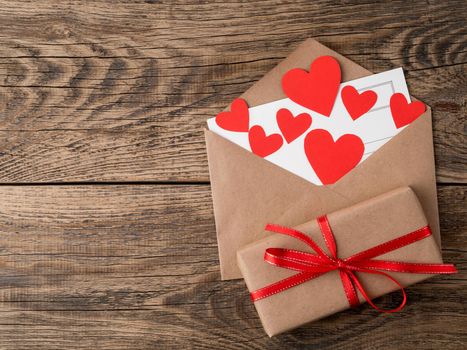card and red hearts in open envelope from brown Kraft paper. Gift box with red ribbon on wooden aged vintage background. Greetings with Valentine's day with copy space for your text