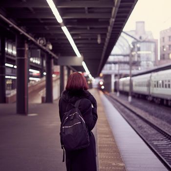 Railway station. Beautiful girl is standing on platform and waiting for train. Woman travels light in evening. Middle-aged lady in warm clothes and coat stands with her back