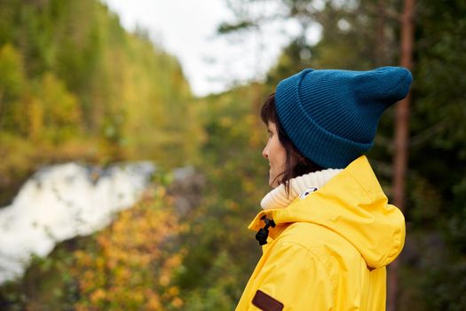 One with nature. Back view of unrecognizable woman looking to forest and waterfall, female in bright yellow jacket and blue hat on on autumn day. Mood Scandinavian weather