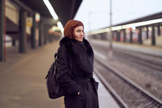 Railway station. Beautiful girl is standing on platform and waiting for train. Woman travels light in evening. Middle-aged lady in warm clothes, coat, in winter. Banner