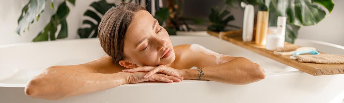 Dreamy young woman leaning on a bathtub side, relaxing with eyes closed at home. Wellness, beauty and care concept