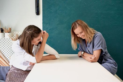 Two beautiful women laugh. Female young businesswoman sit at table in workplace and talk, have fun and gossip