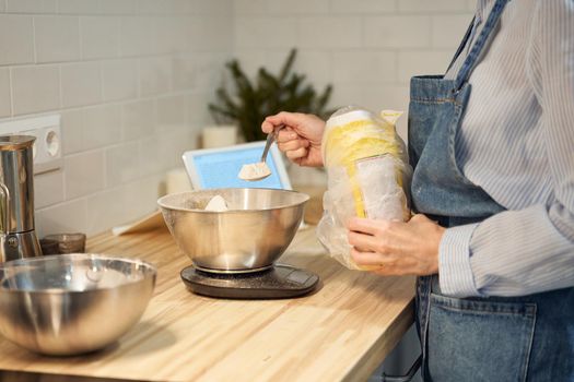 Faceless woman cooking and baking dough on kitchen table at home, apartment, flour, scales, bowls, digital tablet with recipes on table. Homemade food