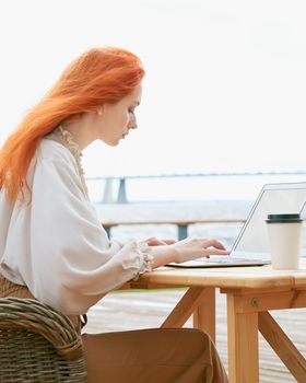 Freelancer woman sitting in cafe on street and remotely working on laptop. Redhaired businesswoman typing on computer