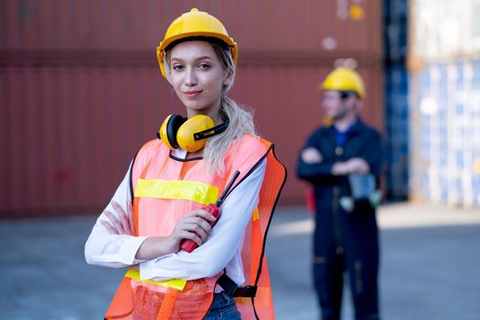 Foreman woman worker stand with confidence by arm-crossed and look at camera with his cargo container co-worker stand in background in workplace area.