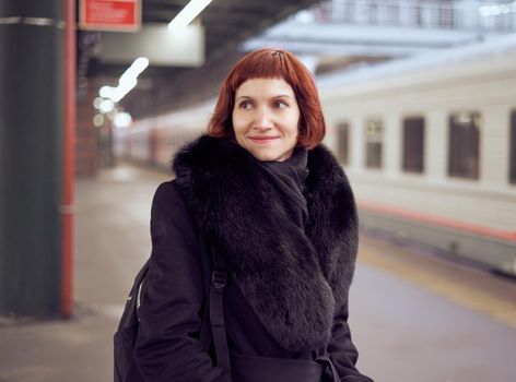 Railway station. Beautiful girl is standing on platform and waiting for train. Woman travels light in evening. Close up portrait of middle-aged lady in warm clothes, coat, in winter