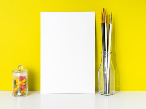 Mockup with clean white canvas, candy in jar, brushes on bright yellow background. Concept for creativity, drawing.