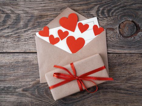 card and red hearts in open envelope from brown Kraft paper. Gift box with red ribbon on wooden aged vintage background. Greetings with Valentine's day